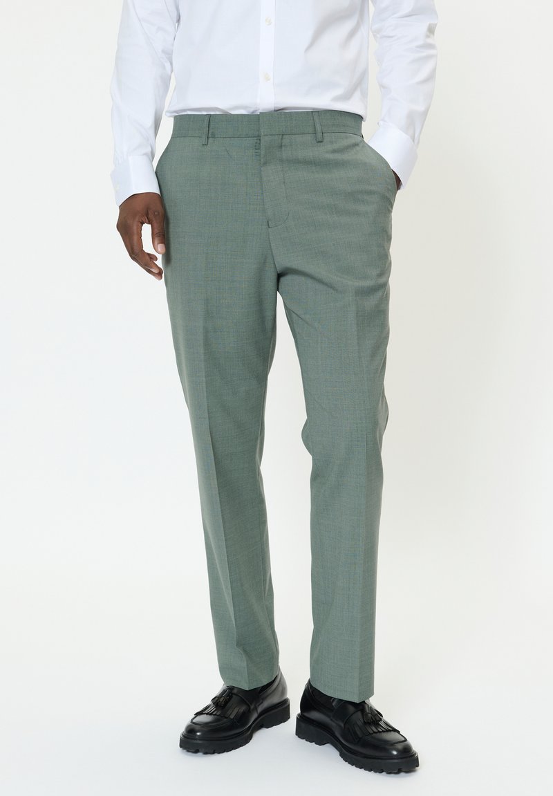 Pantalon vert clair ajusté avec un motif texturé, doté de poches latérales et d'un devant plat, associé à des chaussures noires.