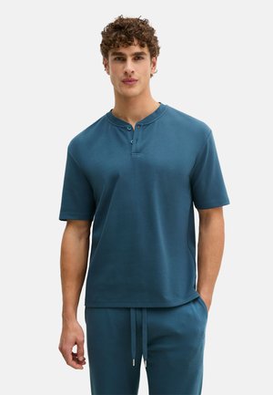 Mann trägt ein türkisfarbenes Henley-Shirt mit kurzen Ärmeln und passende Jogginghosen mit Kordelzug vor weißem Hintergrund.