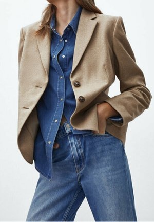 Person wearing a beige blazer over a blue denim button-up shirt tucked into high-waisted blue jeans with one hand in pocket.