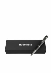 Stylo noir avec accents argentés reposant sur une boîte noire mate, avec "HUGO BOSS" en texte blanc. Design élégant et moderne.