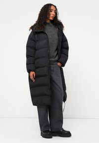 Casaco puffer longo e preto com capuz, apresentando seções acolchoadas, fecho éclair frontal e punhos elásticos. Usado sobre um suéter cinza e calças escuras.