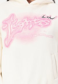 Felpa off-white con grafica testuale rosa asimmetrica, "Lights," e "Low Lights Studios" in una tonalità più chiara; presenta una tasca frontale.