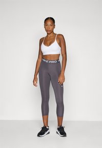 Grijze Nike Pro leggings met een hoge tailleband, gedragen met een witte sportbeha. De outfit heeft een gladde textuur en minimale merkidentificatie.