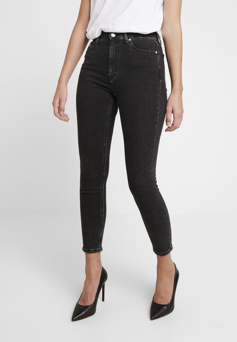 Jeans skinnies negros de cintura alta con una textura de denim suave, detalles de costura prominentes y una silueta ajustada, combinados con tacones negros de punta.