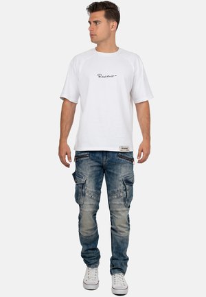 Junger Mann, der nach links schaut, trägt ein weißes T-Shirt mit kurzen Ärmeln und Schriftzug sowie abgenutzte blaue Cargohosen mit weißen Turnschuhen.