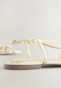Sandali metallici gold con fasce sottili, fibbia decorativa e suola piatta; finitura testurizzata con un motivo screpolato, su uno sfondo chiaro.