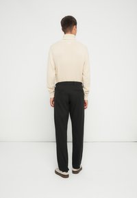 Beige strikket sweater med høj krave, parret med sorte bukser. Outfittet har en afslappet pasform og neutrale farver, båret med lyse sko.