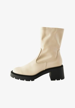 Beige bőr bokacsizma sima felülettel, oldalsó kivágással és vastag fekete sarokkal. Kerek orral és kontrasztos varrással rendelkezik.