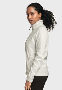 Giacca in pile grigia chiara con colletto alto, chiusura con zip e cuciture laterali. Presenta una superficie liscia e un design aderente.