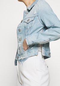 Veste en denim bleu clair avec des détails déchirés, quincaillerie argentée et poches zippées. Présente un design court et un devant boutonné.