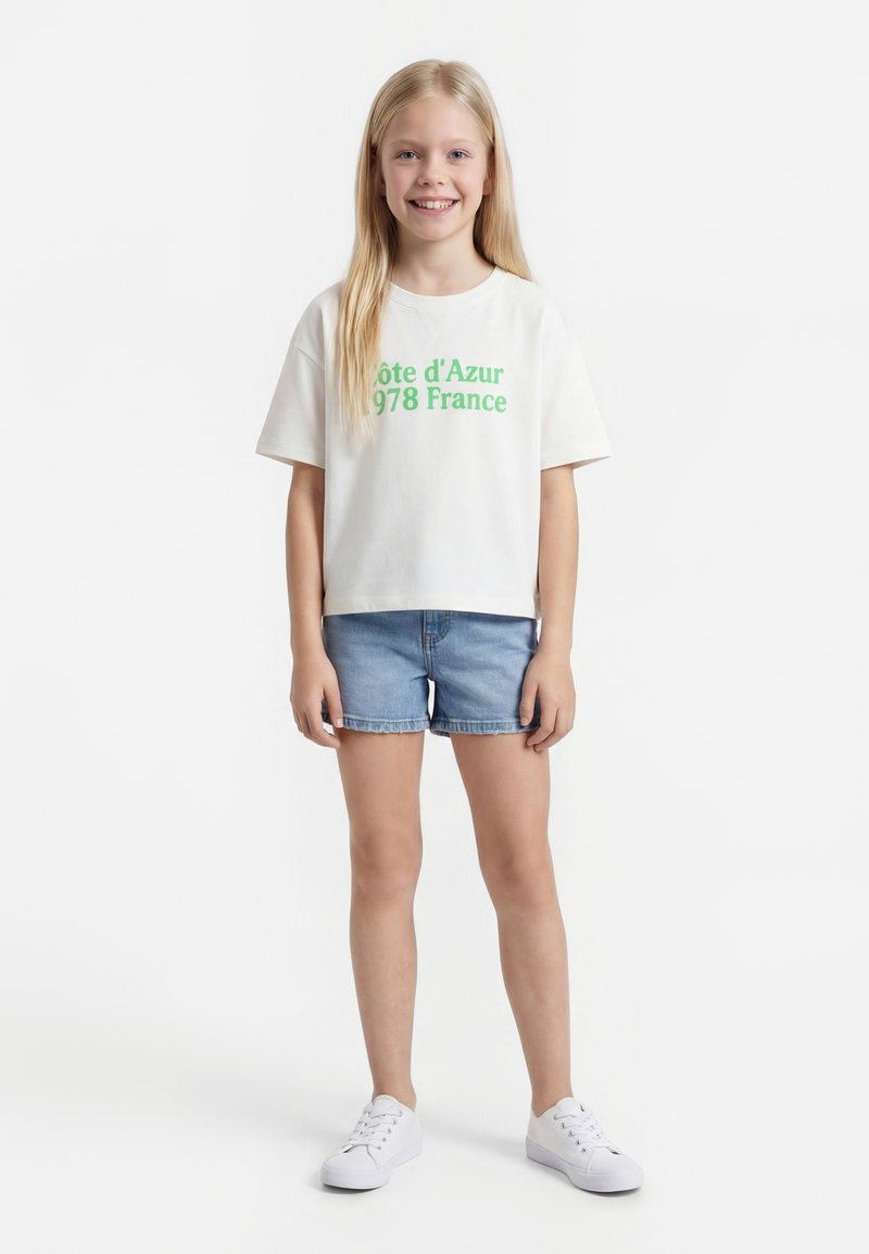 Mladi djevojka stoji i smiješi se, nosi bijelu majicu sa zelenim tekstom, traper kratke hlače i bijele tenisice na jednobojnoj pozadini.