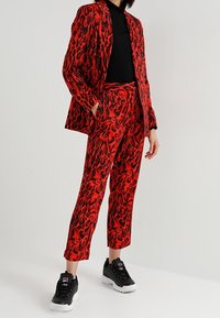 Punainen leopardikuvioinen puku, jossa on tyköistuva bleiseri ja lyhyt housut, yhdistetty mustiin lenkkareihin. Materiaali vaikuttaa sileältä ja hieman kiiltävältä.