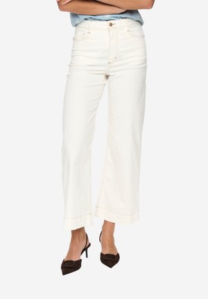 Persona che indossa pantaloni bianchi a vita alta e gamba larga abbinati a décolleté slingback neri con punta appuntita, con le braccia incrociate sopra la vita.