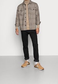 Chemise à carreaux beige, marron et gris avec deux poches poitrine, col roulé beige, jean noir et baskets New Balance marron.