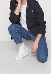 Felpa nera con zip e dettagli rossi e bianchi, t-shirt bianca sotto, jeans blu arrotolati e sneakers bianche con lacci in vista.