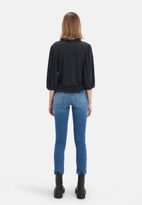 Haut noir à manches longues avec un col à volants, associé à un jean bleu ajusté et des bottines noires. Vue de dos montrant la silhouette de la tenue.