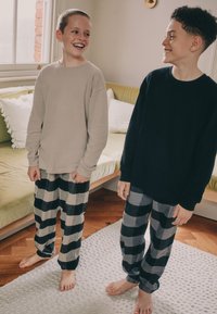 To drenge står på et tekstureret tæppe iført langærmede skjorter og ternede bukser. Den ene skjorte er beige, den anden marineblå; bukserne har grå og sorte tern.