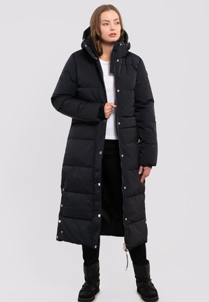 Zwarte pufferjas met een lange snit, capuchon, drukknoop- en ritsafsluitingen, gewatteerde textuur en geribbelde manchetten, gedragen over een witte blouse en zwarte broek.