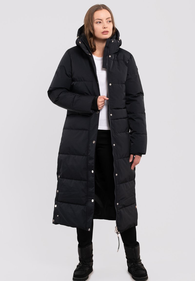 Musta puhvitakki pitkällä vihreällä designilla, hupulla, neppari- ja vetoketjusulkimilla, tikattu pinta ja ribbireunat, päällä valkoinen paita ja mustat housut.