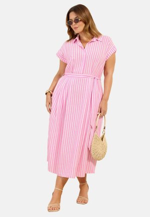 Donna che indossa un vestito midi a righe rosa e bianco con cintura in vita, sandali beige, occhiali da sole rotondi e tiene una borsa intrecciata beige.