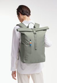 Zaino a sacco in tessuto verde oliva, con una tasca frontale con zip, spallacci regolabili e un piccolo accento in blu e arancione.