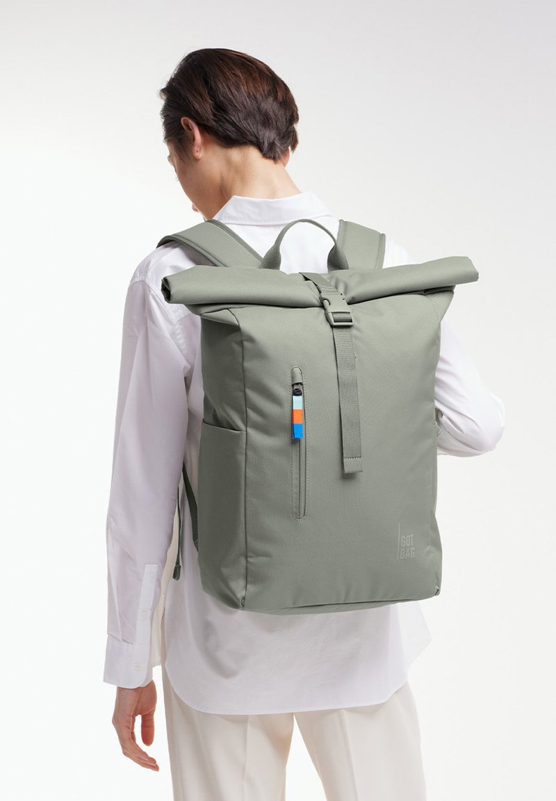 Zaino a sacco in tessuto verde oliva, con una tasca frontale con zip, spallacci regolabili e un piccolo accento in blu e arancione.