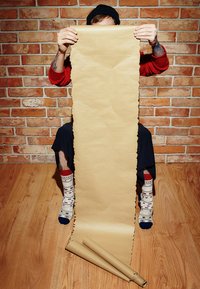 Brązowy krótki papier kraft trzymany w górę, z teksturowanymi brzegami. Otoczenie wokół to ceglana ściana i drewniana podłoga.