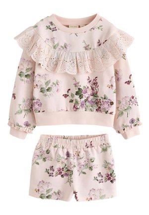 Pige iført en lyserød blomsterlangærmet top med flæsedetaljer og matchende shorts, hvide sokker og hvide sneakers, stående mod en ensfarvet baggrund.