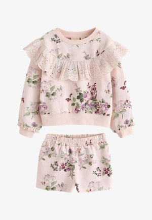 Mädchen trägt ein hellrosa, langärmeliges Oberteil mit Blumenmuster und Rüschen-Detail sowie passende Shorts, weiße Socken und weiße Turnschuhe, steht vor einem einfarbigen Hintergrund.