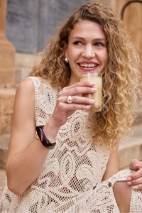 Femme souriante aux cheveux bouclés tenant un verre de latte, portant une robe beige en crochet sans manches et un bracelet foncé, assise en extérieur.