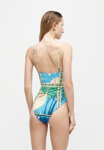Jeune femme aux cheveux mouillés portant un maillot de bain une pièce à motifs bleus et verts, debout de dos sur fond blanc.