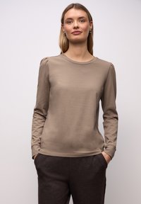Junge Frau mit langen blonden Haaren, die ein beigefarbenes Langarmshirt und dunkelbraune Hosen trägt, steht mit Händen in den Taschen.