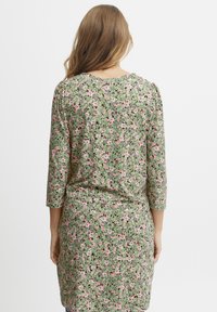 Blumenmusterkleid mit grüner Basis, das mit rosa und weißen Blumen verziert ist. Dreiviertelärmel, runder Ausschnitt und lockere Passform.