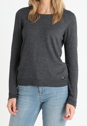 Femme portant un pull à manches longues gris foncé et un jean bleu clair, debout les bras détendus le long du corps.