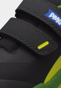 Μαύρο και πράσινο αθλητικό παπούτσι με δύο μαύρα λουράκια με βέλκρο, μπλε και λευκό λογότυπο στο πάνω λουράκι και φωτεινή κίτρινη λεπτομέρεια στο πλάι.