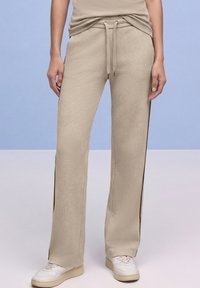 Persona con pantalones de punto ancho beige texturizados con cintura ajustable y zapatillas blancas, de pie frente a un fondo azul claro y blanco.