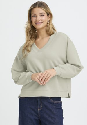 Donna sorridente che indossa una felpa beige chiaro con scollo a V e jeans blu scuro, con le mani incrociate in modo rilassato in vita su uno sfondo semplice.