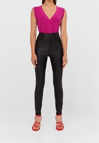 Cardigan sans manches en tricot torsadé rose vif avec fermeture à bouton, associé à un pantalon taille haute en simili cuir noir et des talons rouges à lanières.