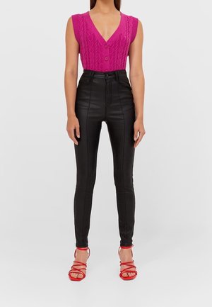 Cardigan sans manches en tricot torsadé rose vif avec fermeture à bouton, associé à un pantalon taille haute en simili cuir noir et des talons rouges à lanières.