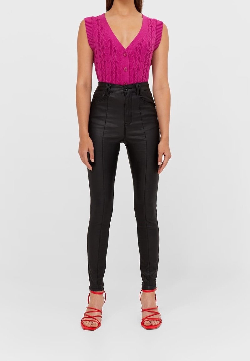 Cardigan sans manches en tricot torsadé rose vif avec fermeture à bouton, associé à un pantalon taille haute en simili cuir noir et des talons rouges à lanières.