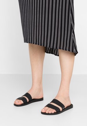 Personne portant des sandales plates tissées noires à double bride et une jupe noire à rayures verticales blanches, debout sur un sol clair uni.