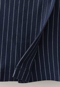 Temno modra tkanina z navpičnimi belimi črtami; ima zložen manšeto, ki razkriva podrobnosti notranjega šivov in gladko teksturo.