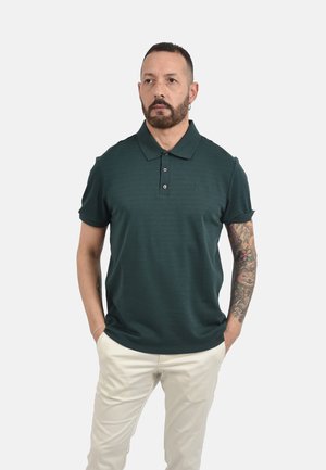 Uomo con capelli corti e barba che indossa una polo verde scuro e pantaloni chiari, mani nelle tasche, mostrando l'avambraccio destro tatuato.