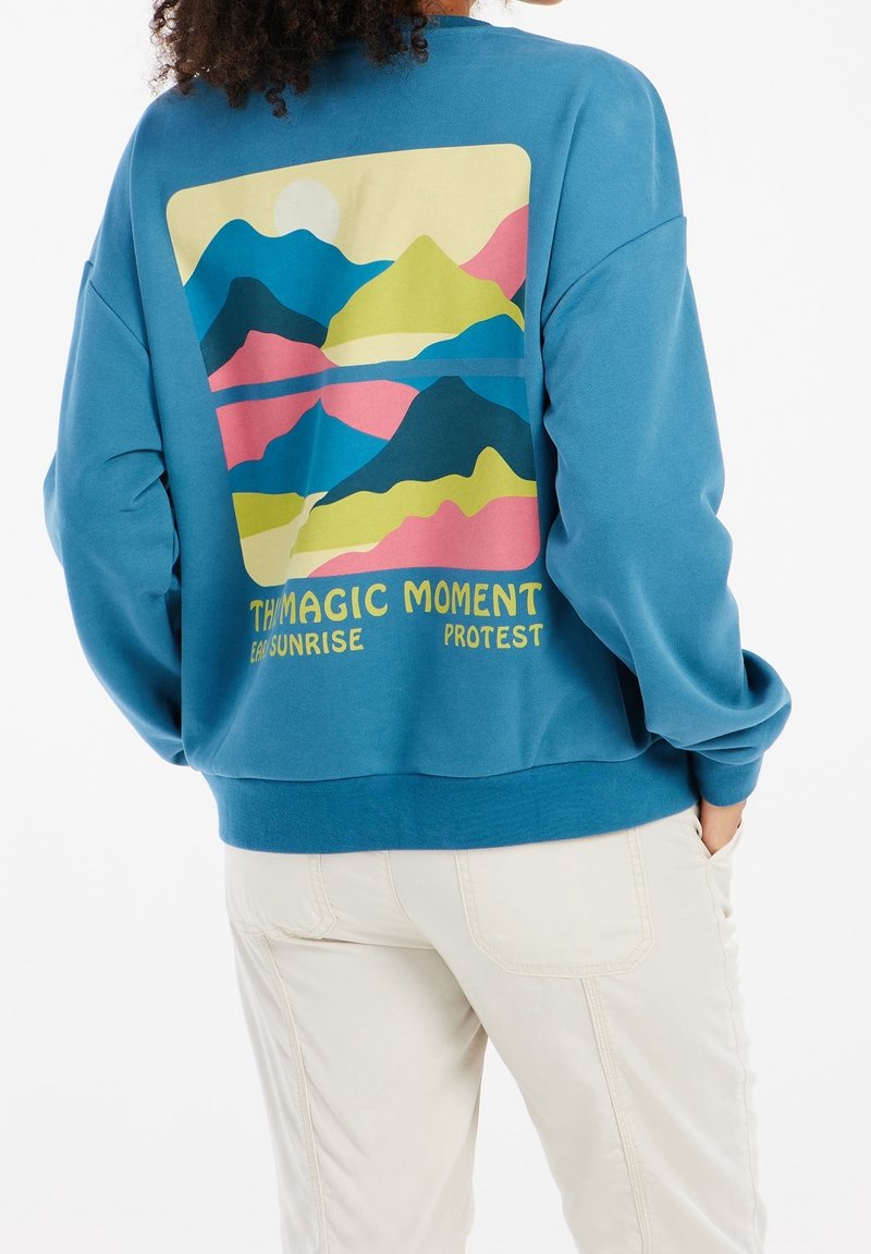 Felpa blu con un design colorato di montagne sul retro, accompagnata dal testo "IL MOMENTO MAGICO PROTESTA DELL'ALBA PREMATURA". Tessuto morbido, vestibilità comoda.