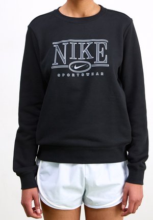 Osoba ubrana w czarną bluzę sportową Nike Sportswear i białe sportowe spodenki, stojąca z ramionami swobodnie opuszczonymi wzdłuż ciała.