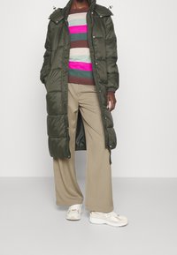 Henkilö pukeutuneena pitkään oliivinvihreään toppatakkiin, raidalliseen neulepaitaan vaaleanpunaisilla ja ruskeilla sävyillä, beigen värisiin leveälahkeisiin housuihin ja valkoisiin New Balance -lenkkareihin.