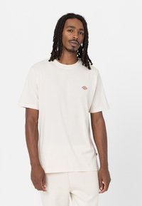 Camiseta de algodón color crema con neckline redondo, mangas cortas y un pequeño logo multicolor en el lado izquierdo del pecho. Diseño sencillo.