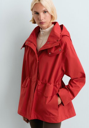 Žena s krátkymi blond vlasmi, ktorá má na sebe červenú kapucňovú bundu cez béžový sveter so zipsom, stojí na jednoduchom svetlosivom pozadí.