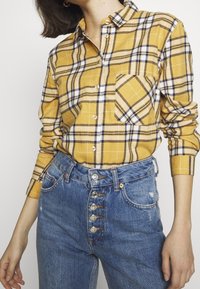 Camicia a quadri gialla con colletto button-down, dotata di taschino sul petto, abbinata a jeans in denim blu a vita alta con dettagli in bottoni argentati.