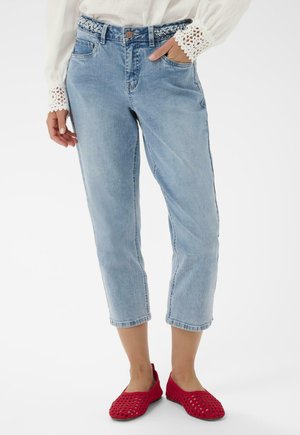 Világoskék farmer rövidített nadrág fehér blúzzal, csipkés mandzsettával, fonott övvel és piros szövött belebújós cipővel viselve.